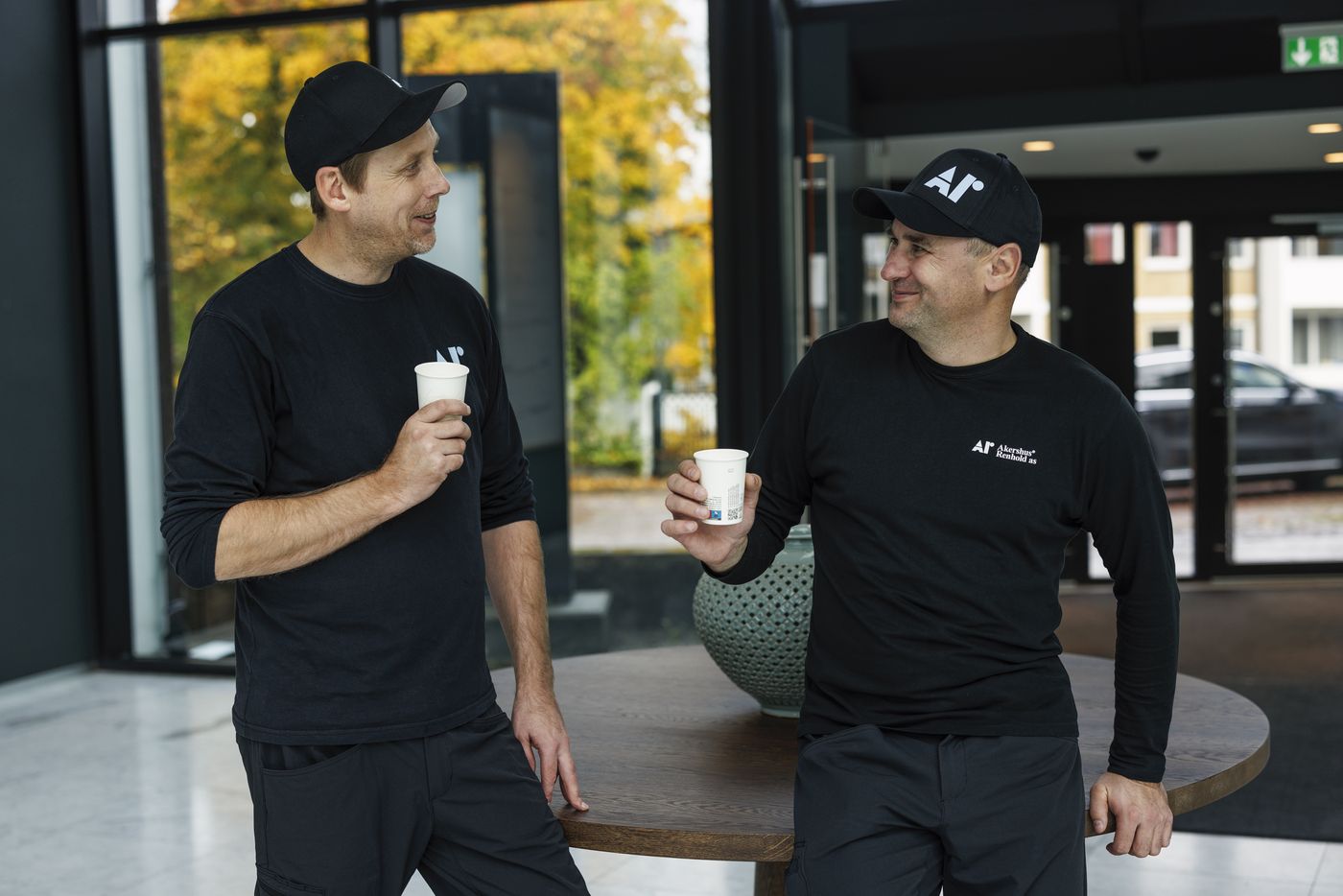 To ansatte i Akershus Renhold-uniform tar en kaffepause i en lys resepsjon.